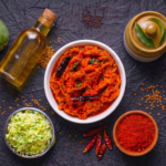 Assorted Indian condiments and spices surrounding a bowl of spicy red pickle on a dark surface, with mangoes and an oil bottle nearby.