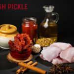 Assorted ingredients for fish pickle: sliced raw fish, jars of red pickled mixture, oil, garlic, mustard seeds, cinnamon sticks, star anise, and spices on a dark surface.