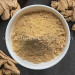 Top-down view of a white bowl filled with light brown powdery substance, surrounded by dried roots on a dark surface.