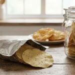 Open bag of tortilla chips on a wooden table, with a jar of chips and a plate in the background to the right.