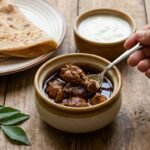 Hand using a spoon to scoop braised meat from a small ceramic pot, with flatbreads on a plate and a yogurt bowl in the background on a wooden table.