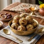 Brass bowl of sesame-coated laddus stacked on a wooden table, surrounded by bowls of nuts and dried fruits.